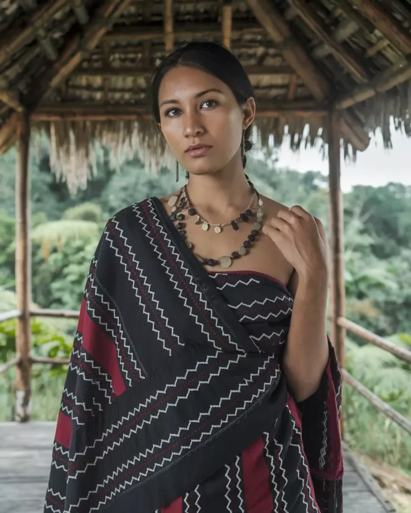 Prompts to Create Beautiful Indian Girls in Diverse Indian Attires AI-generated image of a beautiful Indian girl wearing a traditional black and maroon striped Puan from Mizoram, accessorized with beaded jewelry and standing in a rustic wooden hut, highlighting the simplicity and heritage of Northeast Indian attire by AI Artz.