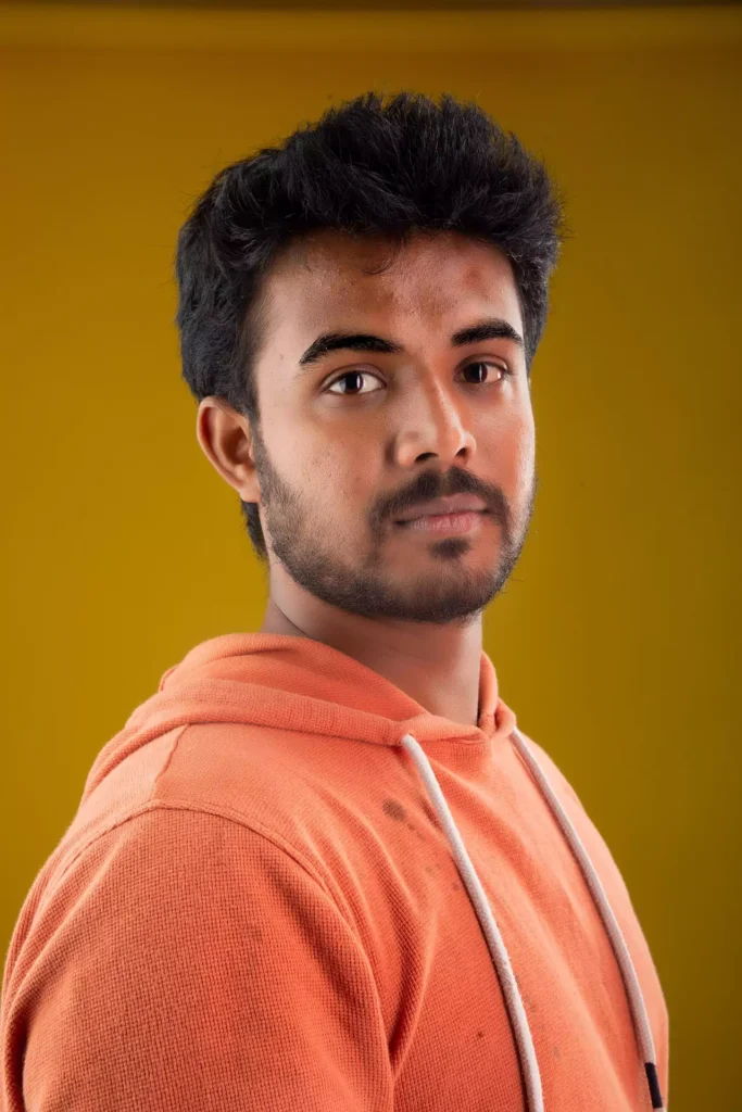 Original reference image of a young Indian man in an orange hoodie against a vibrant yellow background, used for Google Gemini AI photo editing prompts for boys to create stylish, realistic portrait edits and social media-ready looks with modern, trending fashion by AI Artz