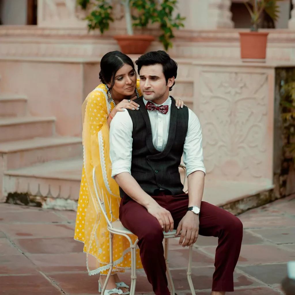 Real Indian couple posing for a romantic outdoor portrait, woman in bright yellow saree with white polka dots gently embracing man in smart casual attire, captured in a heritage courtyard setting, serving as the original reference for Google Gemini AI photo editing prompts for couples, trending realistic love story edits, and authentic modern Indian romance portraits by AI Artz