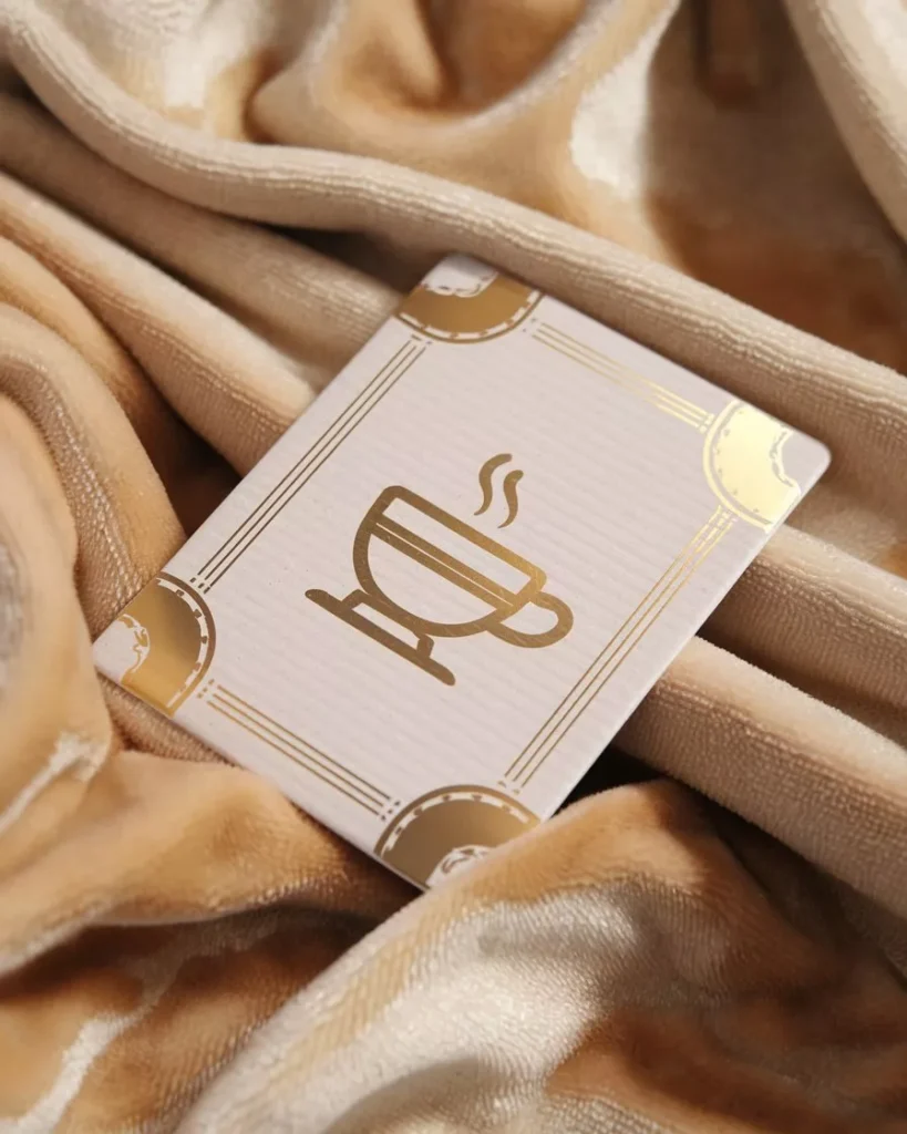 Create Custom AI Images for Coffee Shop Branding AI-generated coffee shop branding image of a Gold-foil Loyalty Card Design coffee cup logo printed on a textured white card with elegant borders, displayed on a soft beige velvet fabric by AI Artz