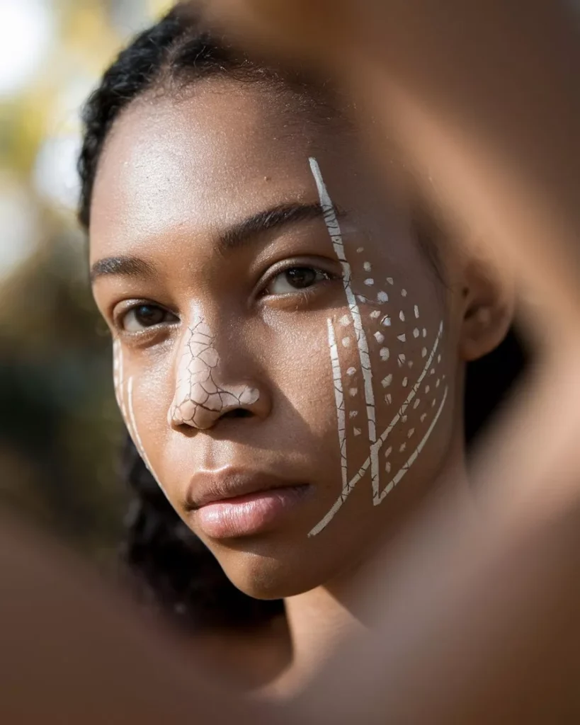 Close up portrait of a woman with dark skin wearing minimalist white face body painting, linear and dotted geometric patterns on cheeks and nose, natural outdoor light, realistic skin texture, high detail reference for AI prompts for body painting by AI Artz