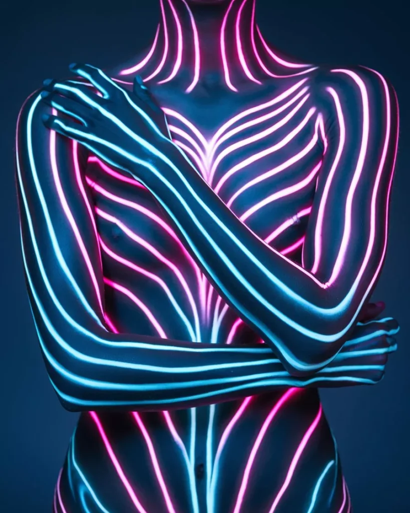 A torso-up shot of a person with symmetrical neon body paint featuring glowing pink and blue organic lines. The model's arms are crossed in a dark, atmospheric studio setting, showcasing hyper-realistic bioluminescent skin art and 2026 neon festival design trends by AI Artz