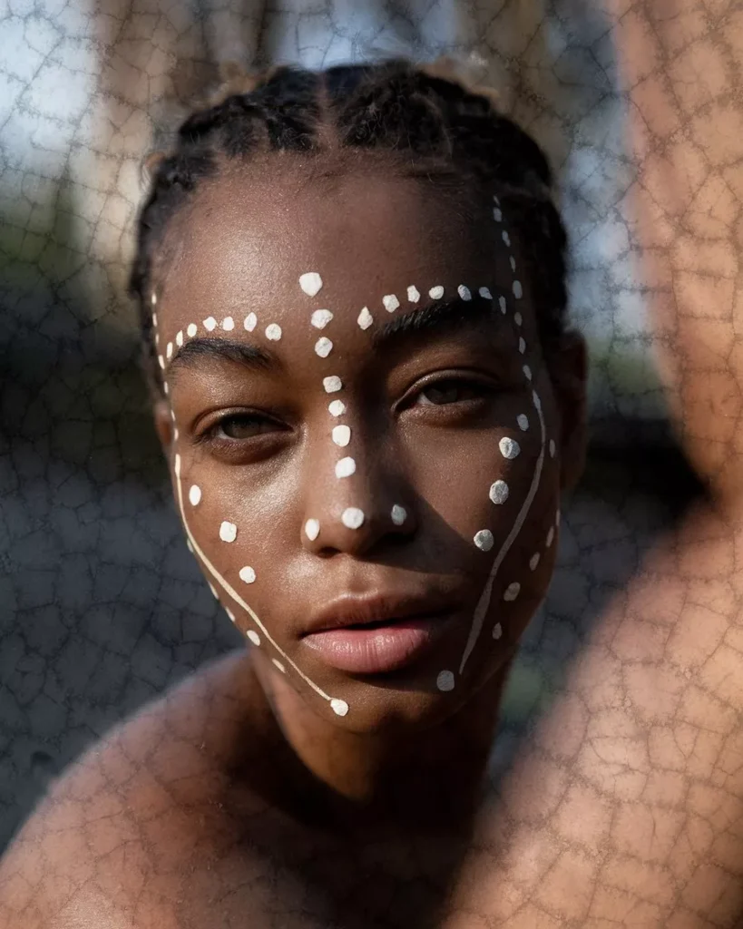Portrait of a woman with dark skin featuring traditional white dotted face body painting, symmetrical tribal-inspired patterns across the forehead cheeks and chin, natural outdoor lighting, realistic skin texture, cultural body art reference for AI prompts for body painting by AI Artz